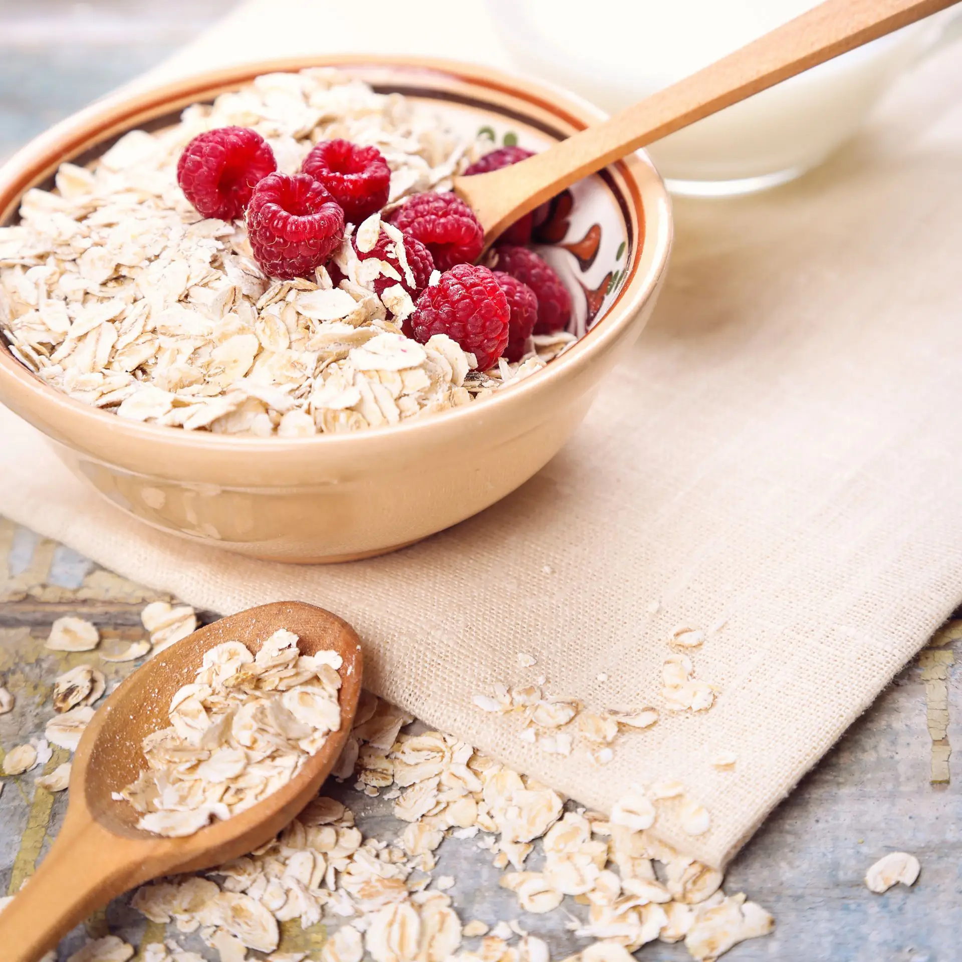 A bowl of oats and raspberries in a bowl and on a spoon on a wooden board with a glass of milk in the background